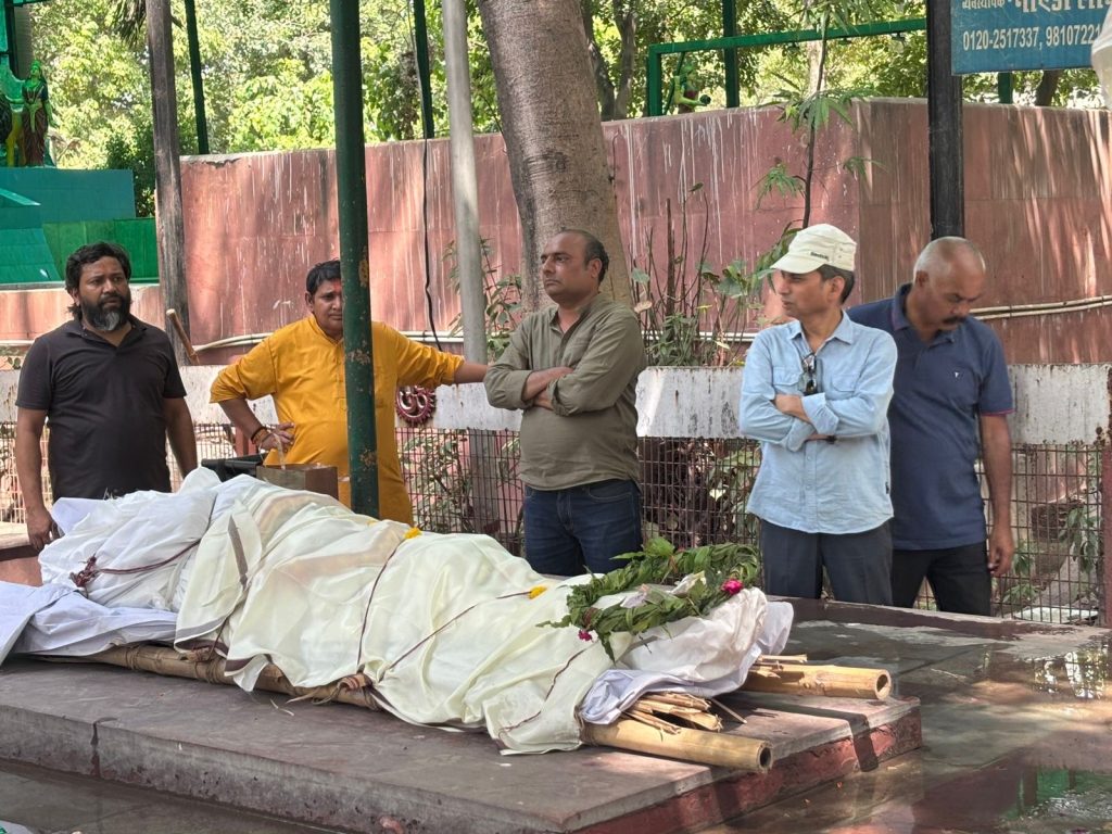 01441300-e2d8-444c-a869-320947270340 | No. 1 Indian Media News Portal Five men stand solemnly beside a body wrapped in white cloth with flowers, laid on a platform outdoors at a cremation site