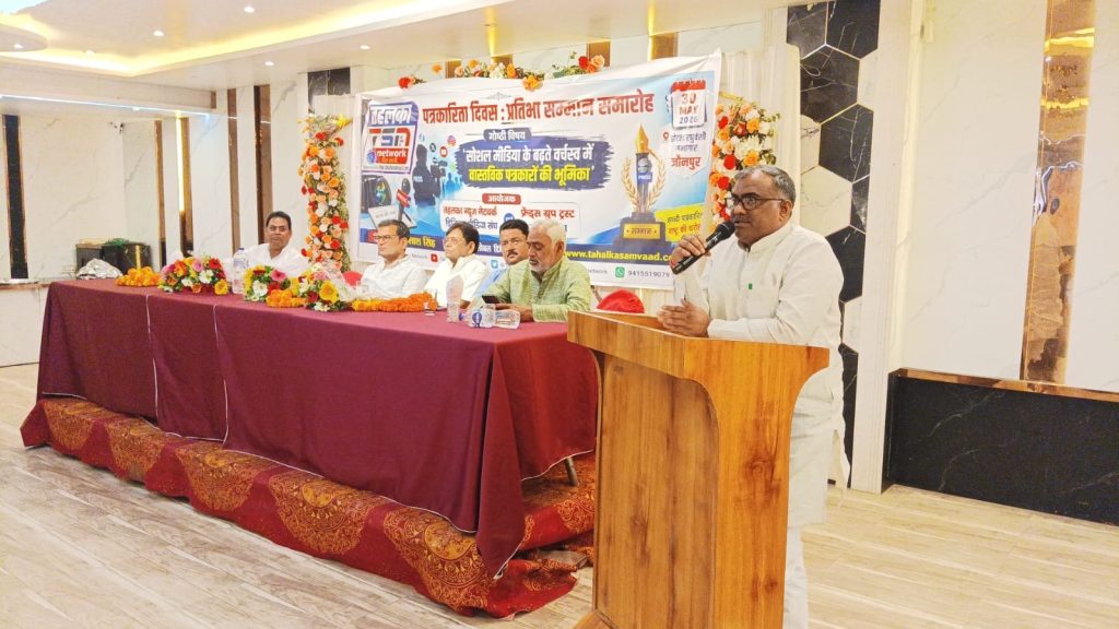 01843136-6ea8-49af-9e23-3d3cc28f1a72 | No. 1 Indian Media News Portal Panel of speakers seated at a long table on a stage, banner behind them, and a man at a wooden podium speaking into a microphone.