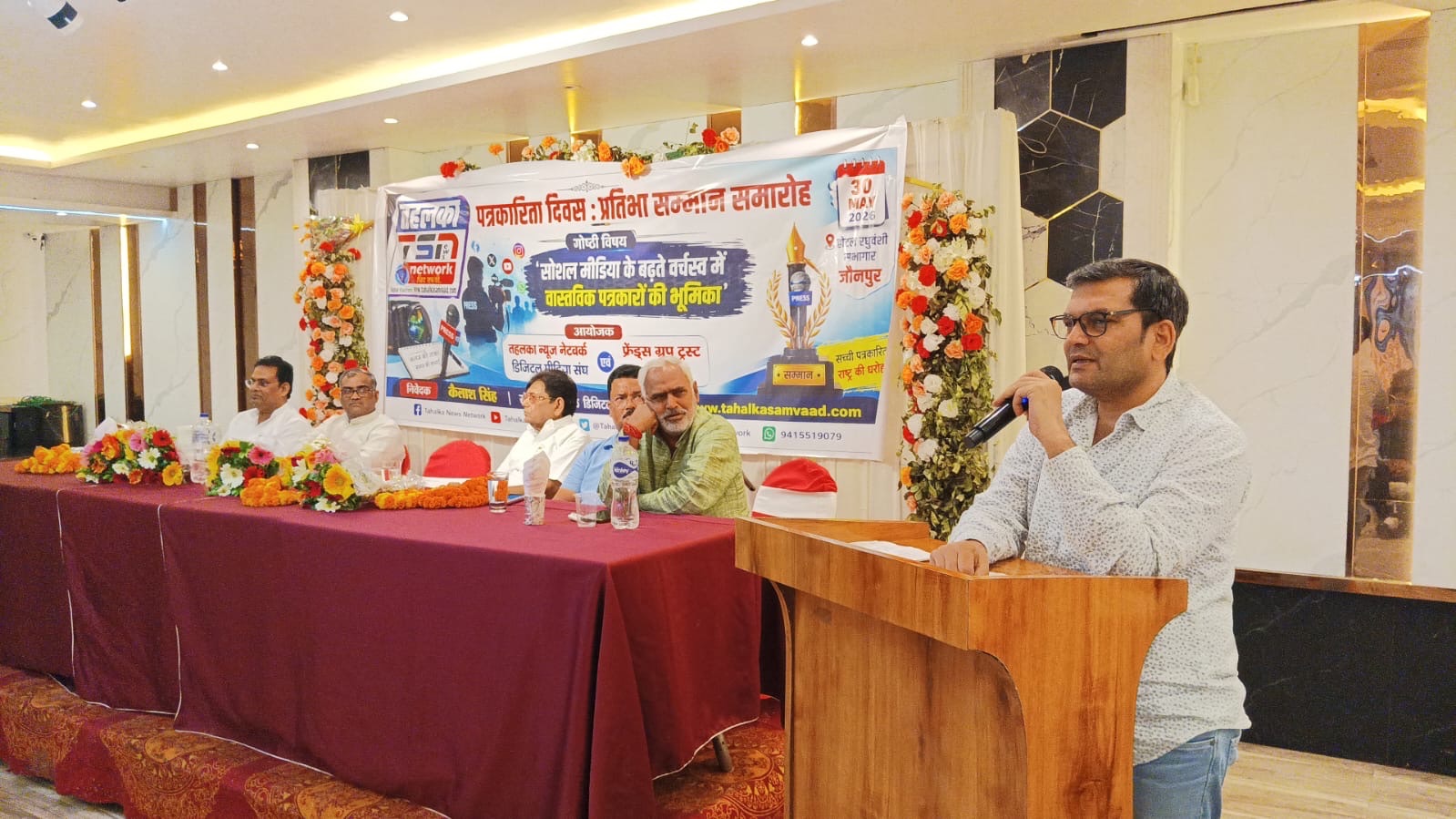 Man speaks into a microphone at a panel discussion, seated beside other panelists with a banner and floral decor in the background.