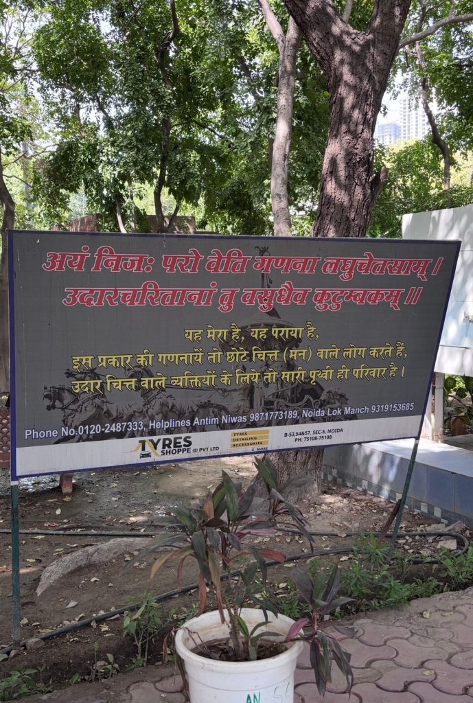 07c4140d-9939-4e8a-b870-5fe37f91998b | No. 1 Indian Media News Portal Large rectangular signboard in a park with Hindi text advertising tyre services; potted plant in the foreground and green trees behind, with a city skyline faintly visible in the background.