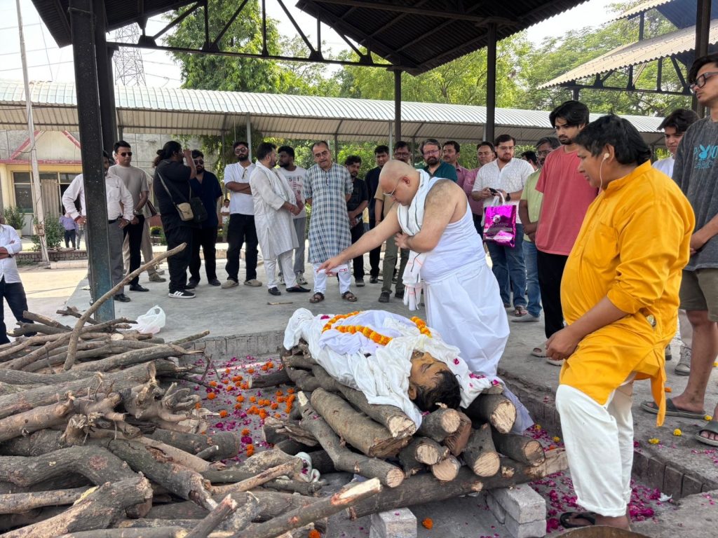 14df97c6-933e-450d-af27-55e801286c53 | No. 1 Indian Media News Portal Man in white performing a Hindu cremation rite beside a body wrapped in white cloth and orange garlands, with mourners gathered nearby.