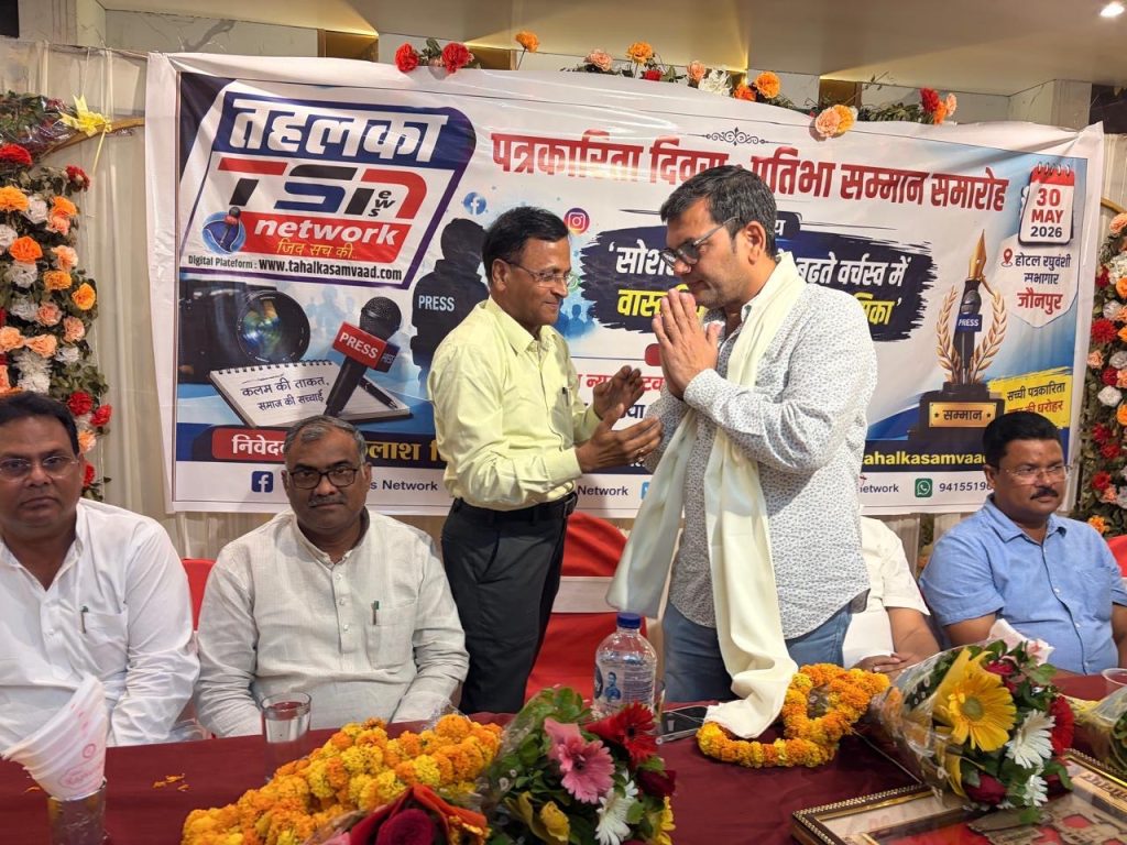 15e57449-1ed8-41dc-994a-62b6890c7efe | No. 1 Indian Media News Portal Two men stand and greet each other with folded hands at a table during a press event, while others sit nearby; a colorful banner for a media network and microphones are in the background, and floral arrangements decorate the scene.