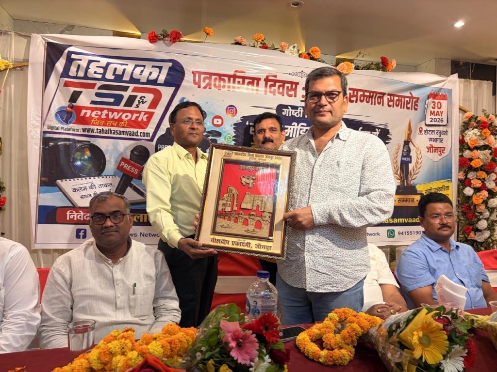 28b96c82-51f1-4fa5-8ced-b042f220d857 | No. 1 Indian Media News Portal Two men stand on a stage during an award ceremony, one handing a framed certificate to the other while flowers and a banner backdrop are visible behind them.