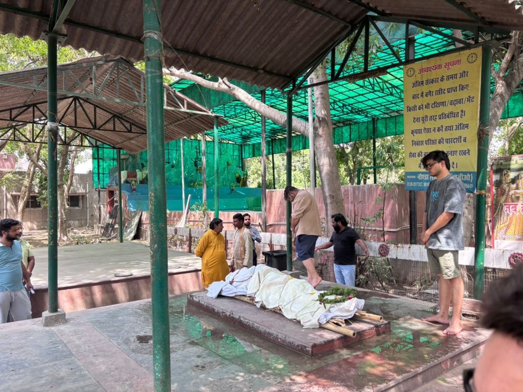 3f257139-12f1-4919-bd06-e6876cc8c409 | No. 1 Indian Media News Portal Public cremation altar under a green canopy; a body wrapped in white cloth lies on a bier as bystanders watch nearby.