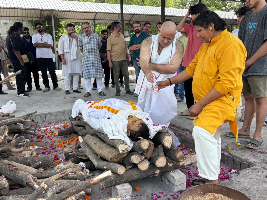 3fbe7277-e00b-4661-8f6a-b73dc38bee4c | No. 1 Indian Media News Portal A group of mourners watch as a body wrapped in white lies on a wood pyre, with orange flower petals scattered nearby for cremation.