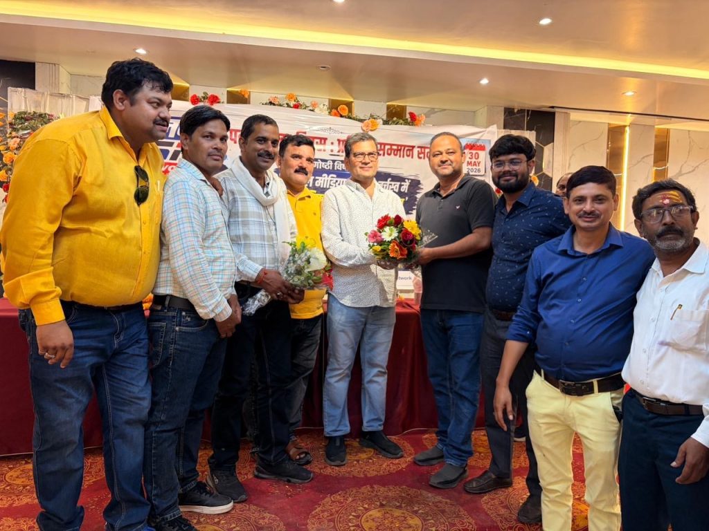533c4987-7cb4-4e55-83ab-9a932382530a | No. 1 Indian Media News Portal Group of men posing for a photo at an indoor event; one man hands a bouquet as others smile, with a decorated banner and flowers in the background.