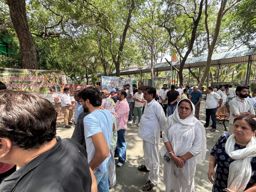 6d696cdd-8591-424a-96f5-21971594a6d5 | No. 1 Indian Media News Portal Group of people gathered in a shaded park for an outdoor event, banners in Hindi visible in the background.
