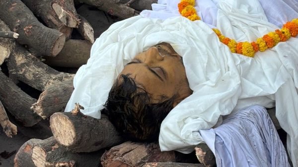 Deceased person wrapped in white shroud lying on stacked firewood, with a marigold garland around the neck."