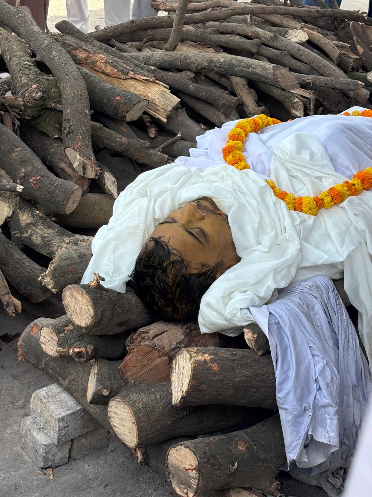 Deceased person wrapped in white shroud lying on stacked firewood, with a marigold garland around the neck."