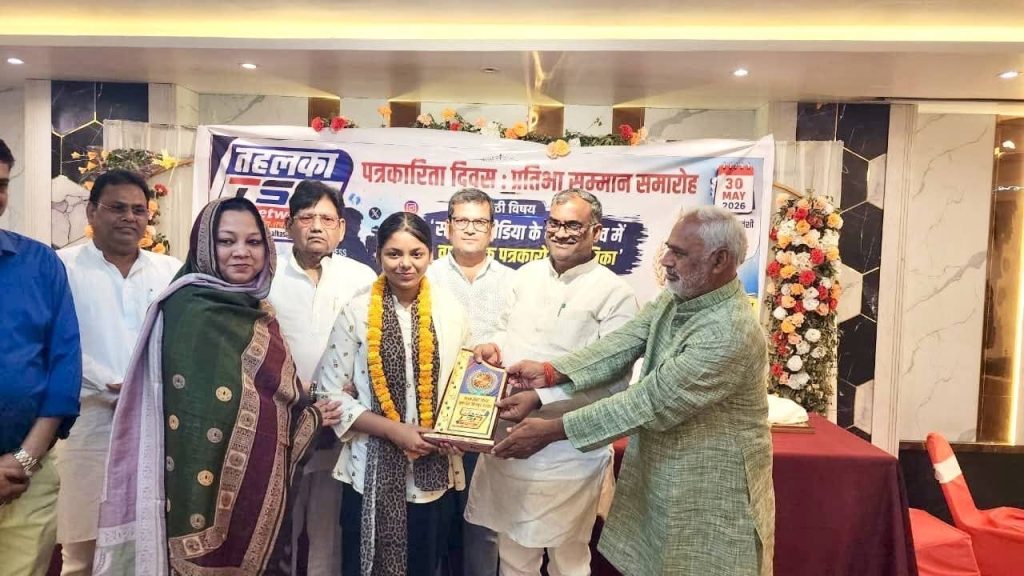 7aab2e12-5cfa-4877-b778-ff1a75b62f5c | No. 1 Indian Media News Portal Group presentation: a plaque is handed to a young woman wearing a garland during an indoor award ceremony, with a banner and flowers in the background.