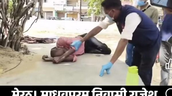 News thumbnail: man lying on pavement while a bystander in gloves offers water as others look on; people outdoors wearing masks in the background.