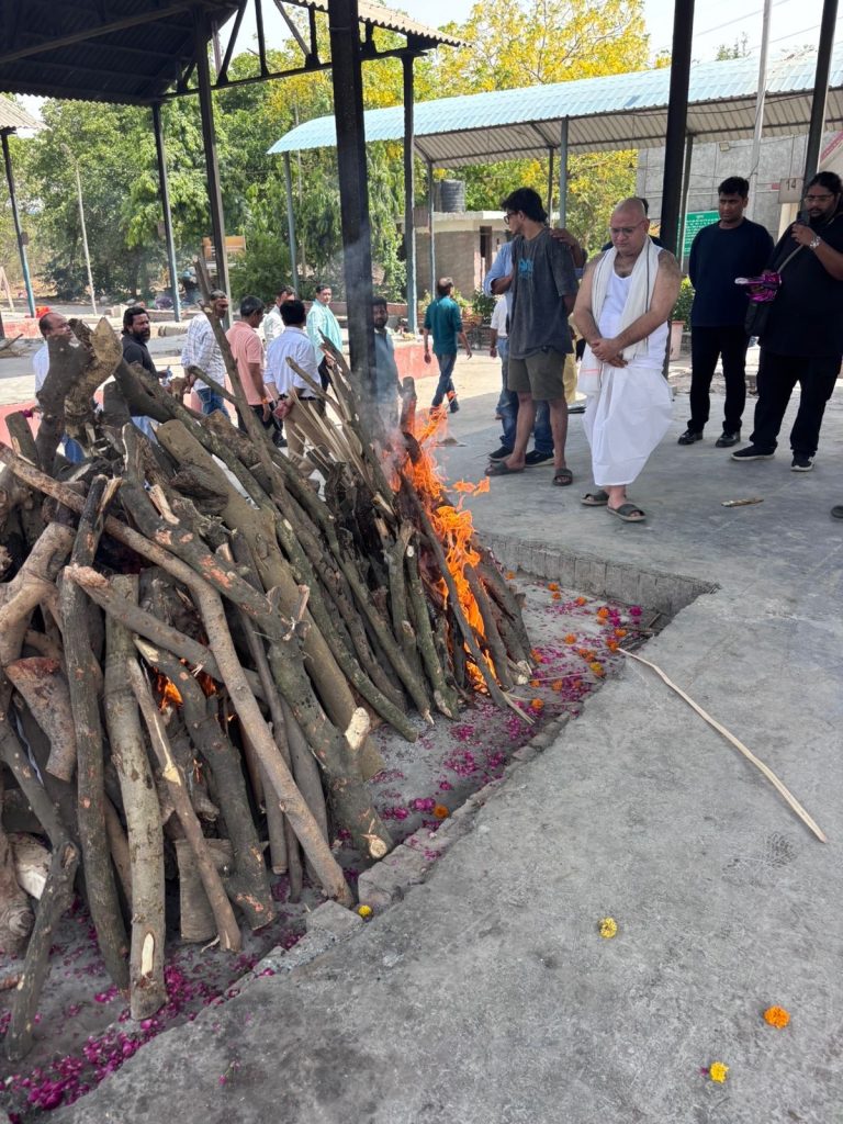 8b181765-8c5e-459c-bee7-af16594a0aef | No. 1 Indian Media News Portal Group of men stand around a large bonfire of stacked wood in an open courtyard, with flower petals scattered nearby.