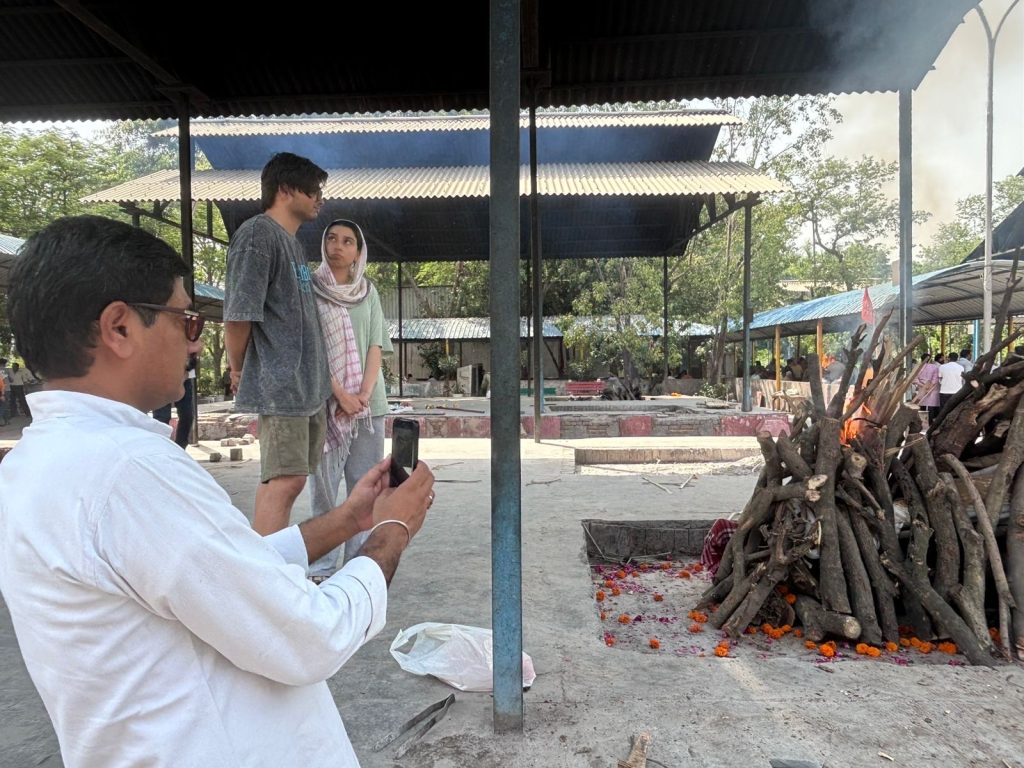 8e545189-ebe6-4724-bb13-0f1ebb25b8b8 | No. 1 Indian Media News Portal Man in a white shirt sits at left, filming with a smartphone while a couple stands nearby under a shaded open shelter; in the foreground a wood pile on fire with orange marigold petals scattered on the ground.