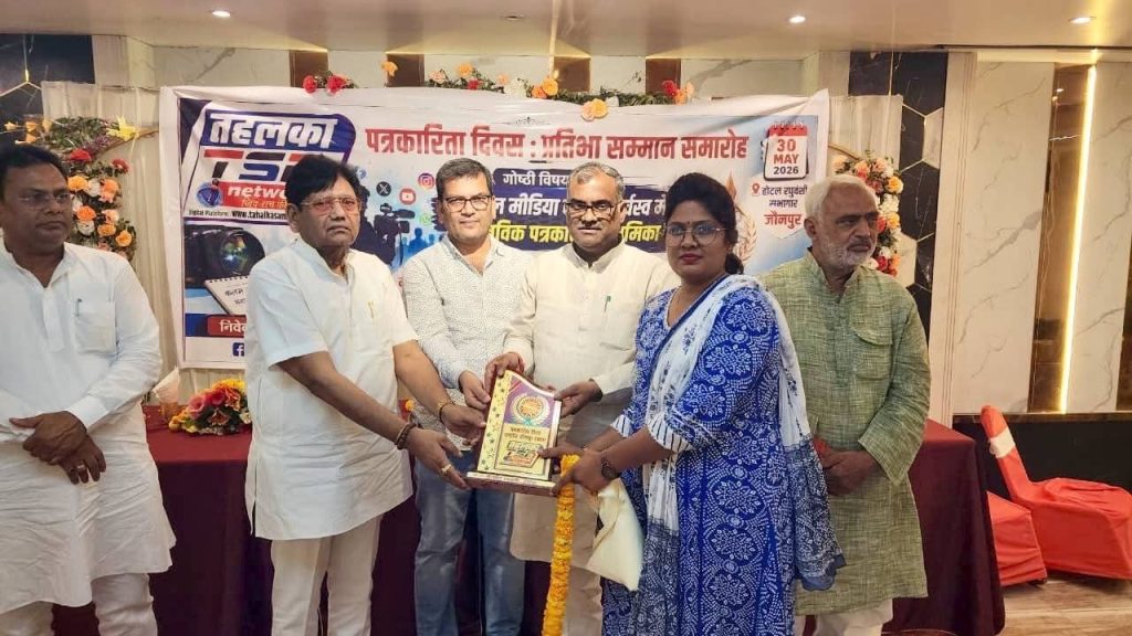 ae5135b9-30b3-42d7-8ea3-9230a5e40ac8 | No. 1 Indian Media News Portal Six people stand on a stage as a woman in blue attire receives a plaque during an award ceremony, with a banner and floral garlands in the background.