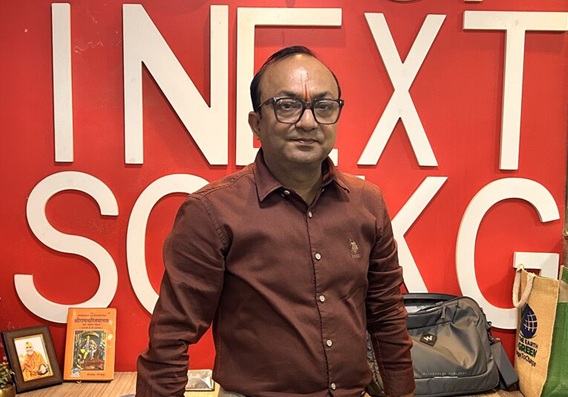 alok tripathi | No. 1 Indian Media News Portal Man in a maroon shirt and glasses stands before a red wall with large white letters; framed photos and a gray bag on a table.