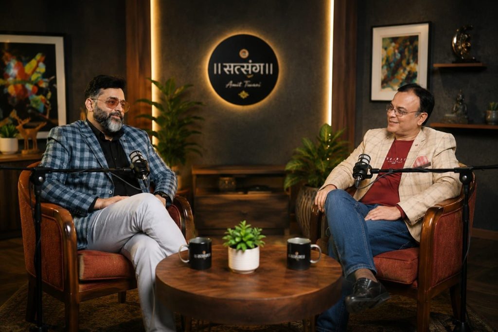 Two men in a podcast studio sit in armchairs across from each other, microphones in front, chatting at a round wooden table with mugs and a small plant in the middle.