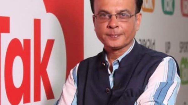 Man with glasses in a blue striped shirt and dark vest, arms crossed, standing in front of a red and white 'Talk' banner.
