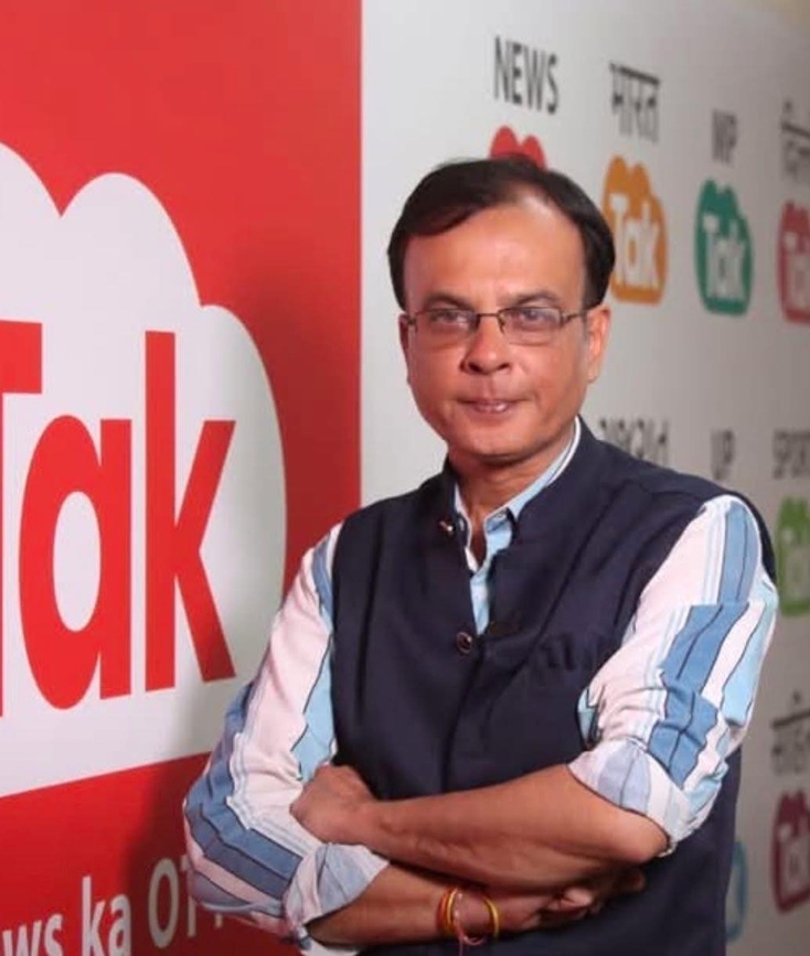 Man with glasses in a blue striped shirt and dark vest, arms crossed, standing in front of a red and white 'Talk' banner.
