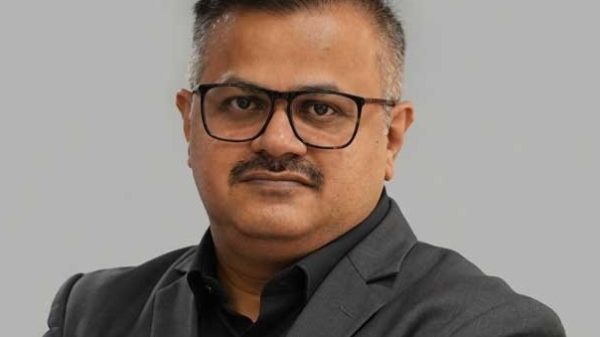 Professional head-and-shoulders portrait of a man wearing black shirt and dark suit with glasses, looking at the camera.