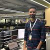 Smiling man wearing a PRESS badge stands at a desk with multiple computer monitors in a newsroom-like office.