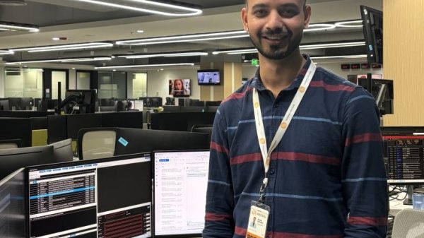 Smiling man wearing a PRESS badge stands at a desk with multiple computer monitors in a newsroom-like office.