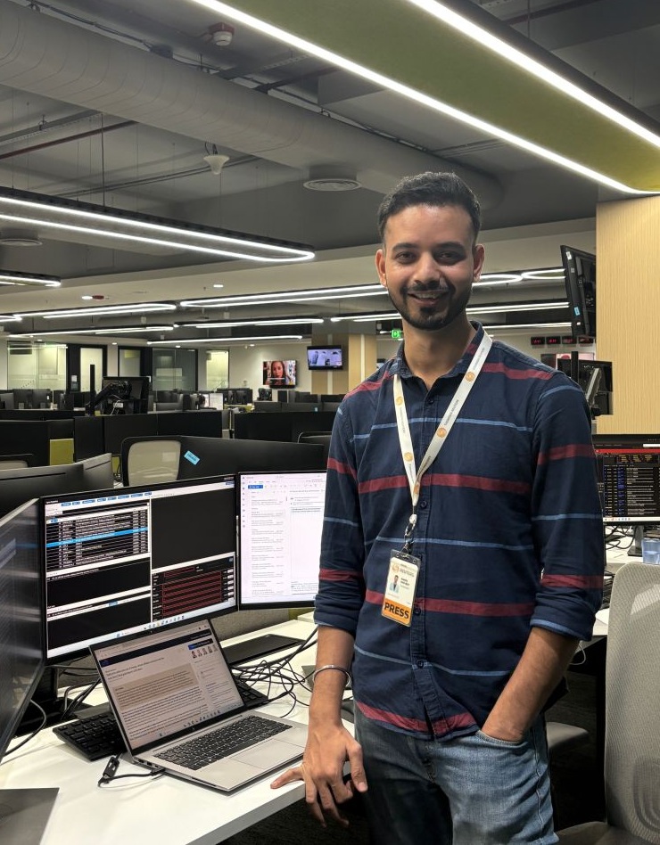 Smiling man wearing a PRESS badge stands at a desk with multiple computer monitors in a newsroom-like office.
