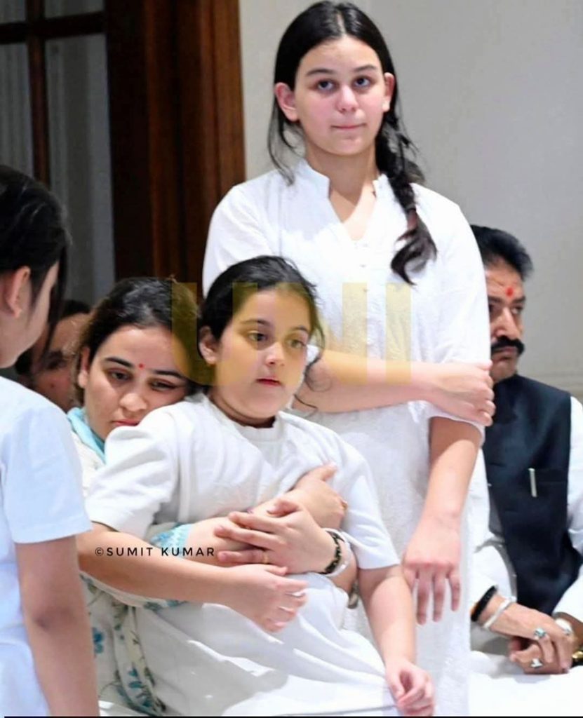 aparna__ | No. 1 Indian Media News Portal Young girl in white being hugged from behind by a woman at a white-clad group gathering, with others seated nearby.