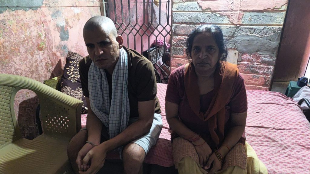 Man and woman sit on a pink bed in a small, worn room with exposed brick walls behind them, looking at the camera.