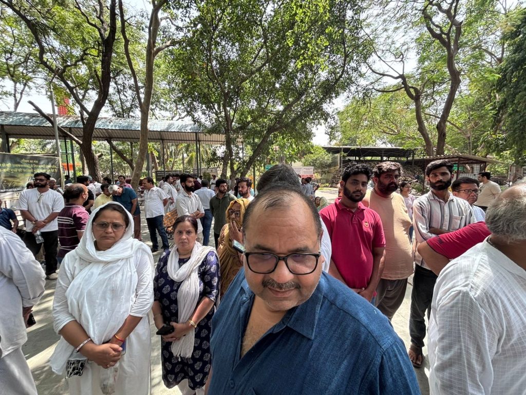 b072f3ac-0451-4846-b2d9-2c7d652362cc | No. 1 Indian Media News Portal A crowd of people standing in a queue outdoors under trees, some looking at the camera, in a sunny park area.