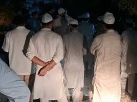 Group of men wearing traditional white garments and skullcaps standing with their backs to the camera at night outside.