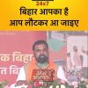 Speaker at a political rally delivering a speech on a decorated stage with a yellow banner reading 'JAN SHRISTI 24x7' in front of him. Behind him, a Hindi banner says 'बिहार आपका है आप लौटकर आ जाइए'.