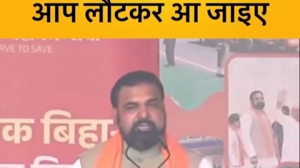 Speaker at a political rally delivering a speech on a decorated stage with a yellow banner reading 'JAN SHRISTI 24x7' in front of him. Behind him, a Hindi banner says 'बिहार आपका है आप लौटकर आ जाइए'.
