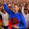 Man in a blue kurta waves to a large crowd while holding a bouquet of red roses at a rally.