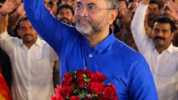 Man in a blue kurta waves to a large crowd while holding a bouquet of red roses at a rally.