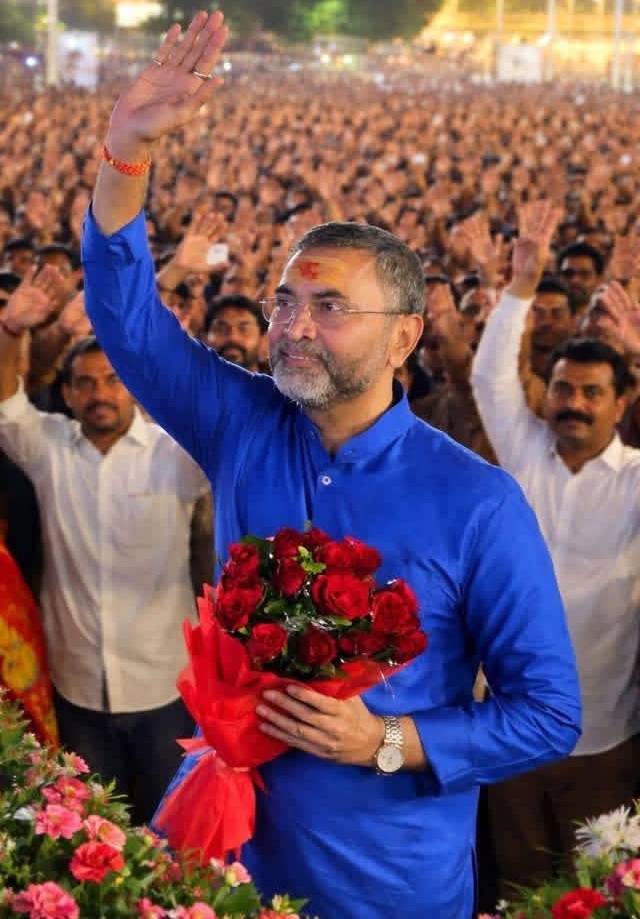 Man in a blue kurta waves to a large crowd while holding a bouquet of red roses at a rally.