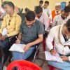 Group of men seated in a hall, several filling out forms on clipboards at a community event.
