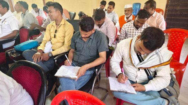 Group of men seated in a hall, several filling out forms on clipboards at a community event.
