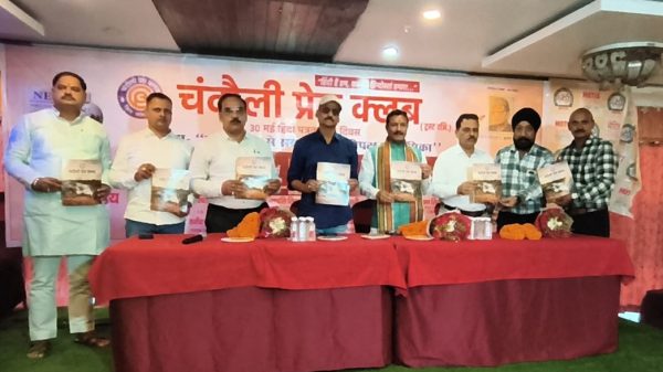 Eight men stand behind a red table on a stage, each holding a copy of a book during a Hindi-language event; a banner with Hindi text is in the background.
