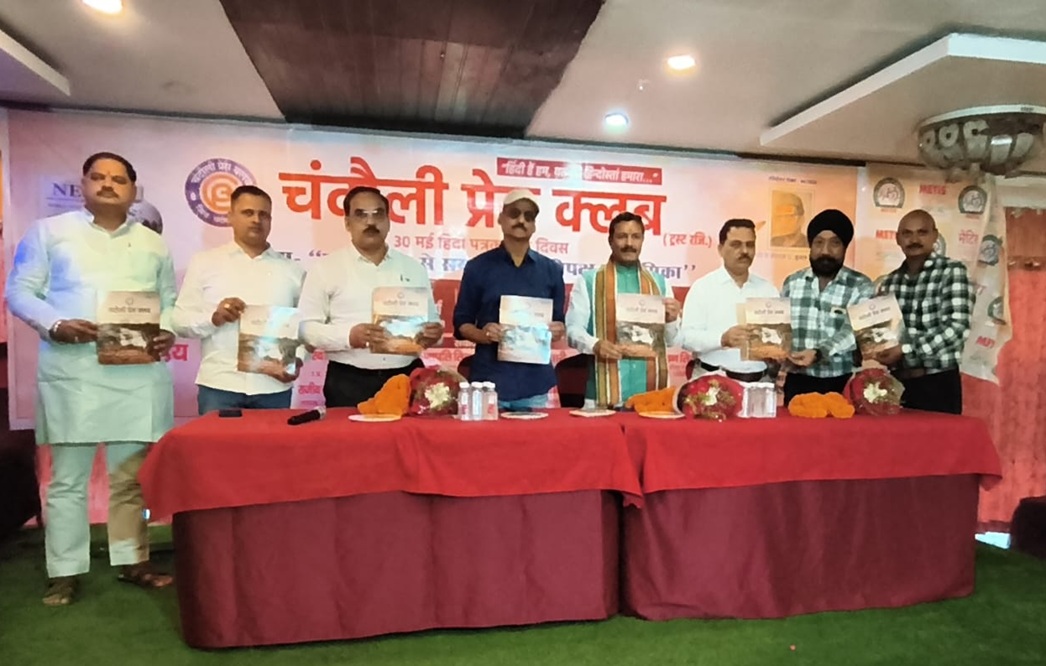Eight men stand behind a red table on a stage, each holding a copy of a book during a Hindi-language event; a banner with Hindi text is in the background.