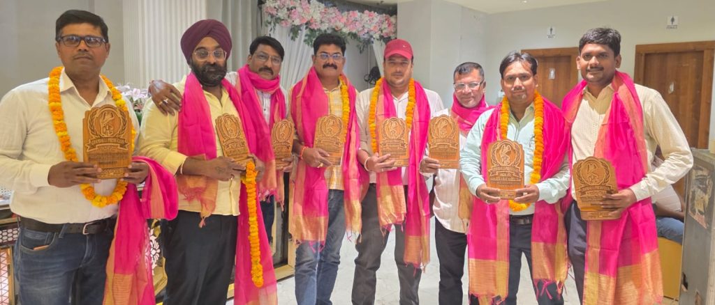chandauli_press | No. 1 Indian Media News Portal Nine men in pink scarves and marigold garlands pose indoors, each holding a carved wooden plaque from an award ceremony.