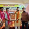 Group of men on a stage, exchanging a framed award with scarves, at a cultural event backdrop written in Hindi.
