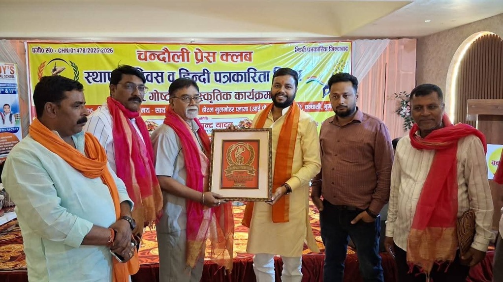 Group of men on a stage, exchanging a framed award with scarves, at a cultural event backdrop written in Hindi.