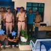 Group of Indian police officers standing behind two seated detainees whose faces are covered, inside a police station room with a desk and plant.