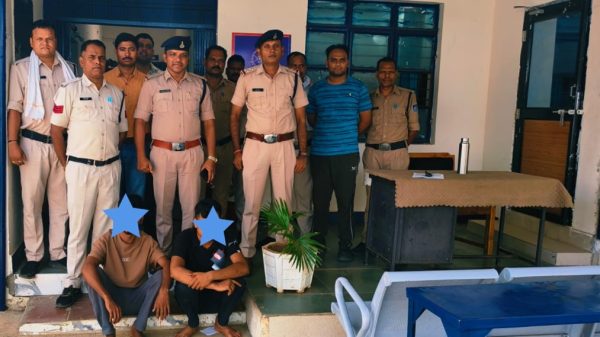 Group of Indian police officers standing behind two seated detainees whose faces are covered, inside a police station room with a desk and plant.