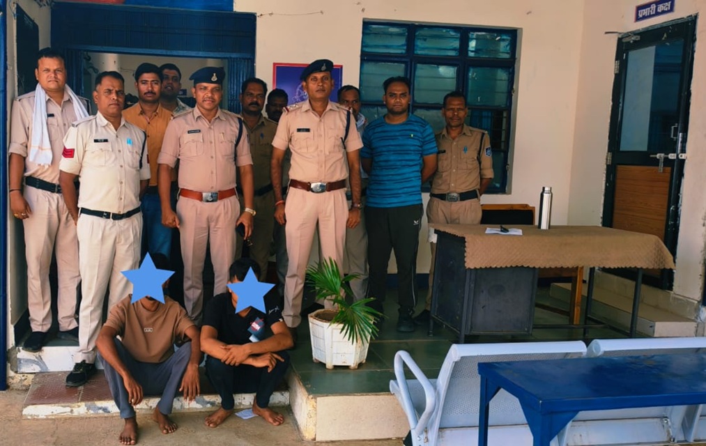 Group of Indian police officers standing behind two seated detainees whose faces are covered, inside a police station room with a desk and plant.