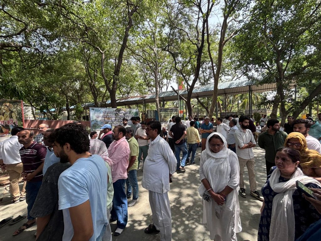 d31322fe-3af0-4bac-9ba5-e1c67b9fc178 | No. 1 Indian Media News Portal People lining up outdoors under trees, with banners/posters in the background.