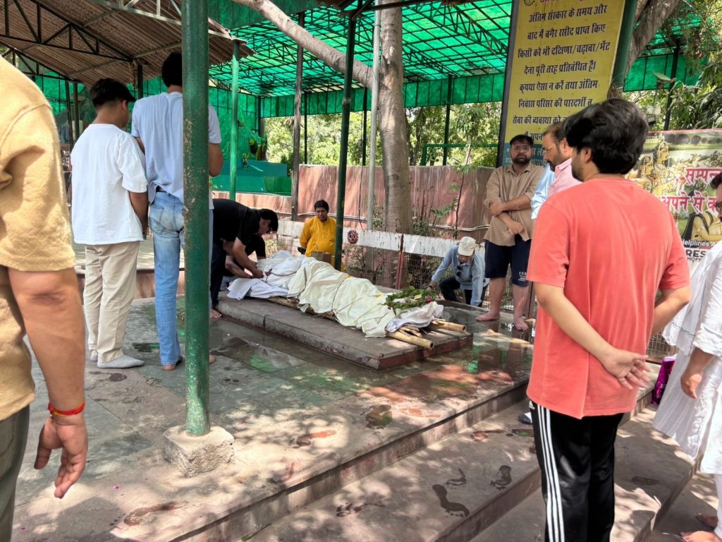 db177ed8-eeec-48c8-bb41-3bcab4434c2c | No. 1 Indian Media News Portal A group of men gathered around a body wrapped in white cloth on a low platform, with flowers, at an outdoor cremation site under a green canopy.