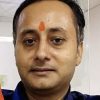 Close-up of a man with an orange tilak on his forehead, wearing a dark blue shirt, indoors.