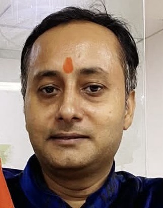 Close-up of a man with an orange tilak on his forehead, wearing a dark blue shirt, indoors.
