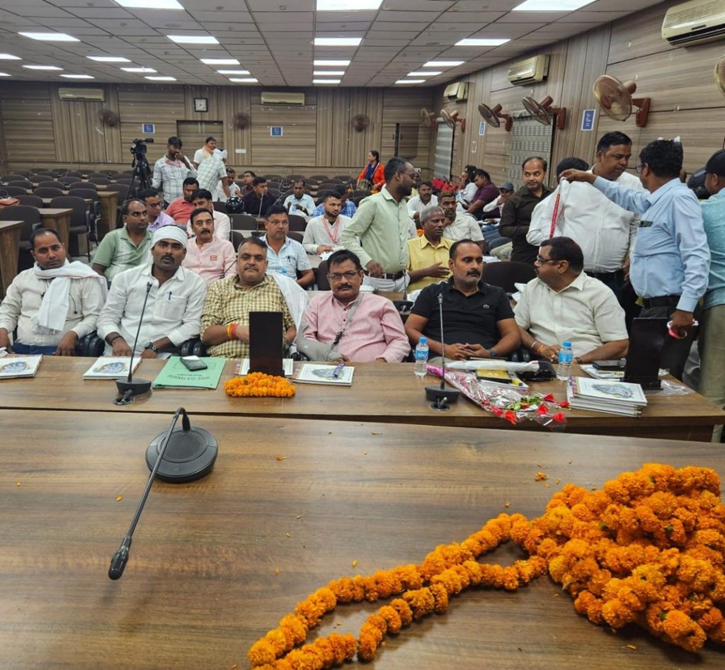ghazipur samman_3 | No. 1 Indian Media News Portal Group of men seated at a long conference table in a hall, with marigold garlands and documents on the table, as others stand and chat in the background.